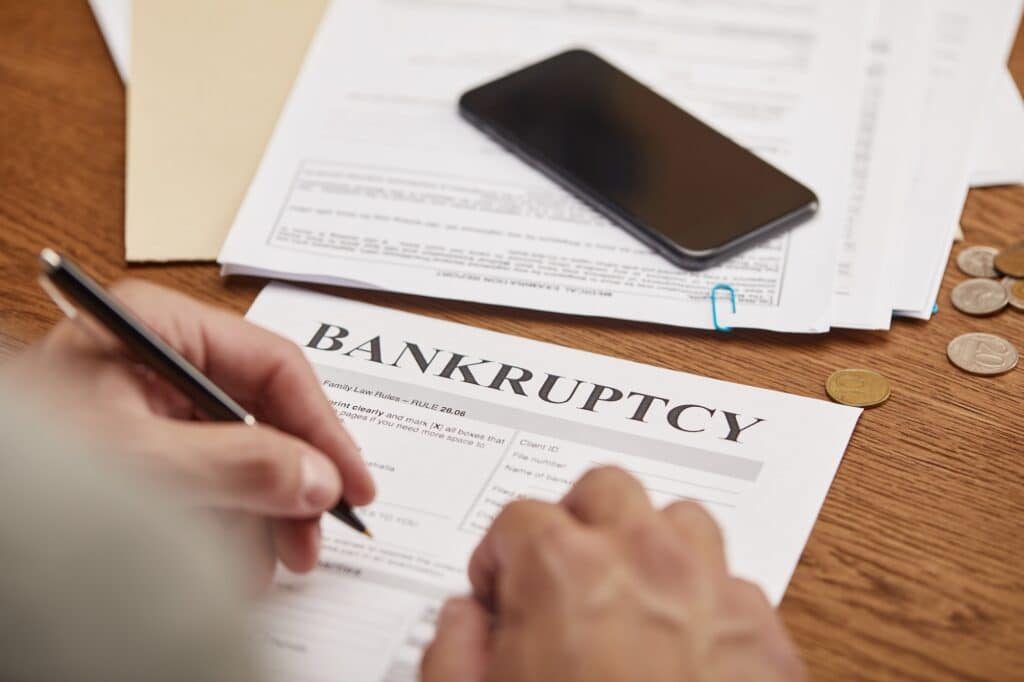 partial view of businessman filling in bankruptcy form at wooden table with smartphone